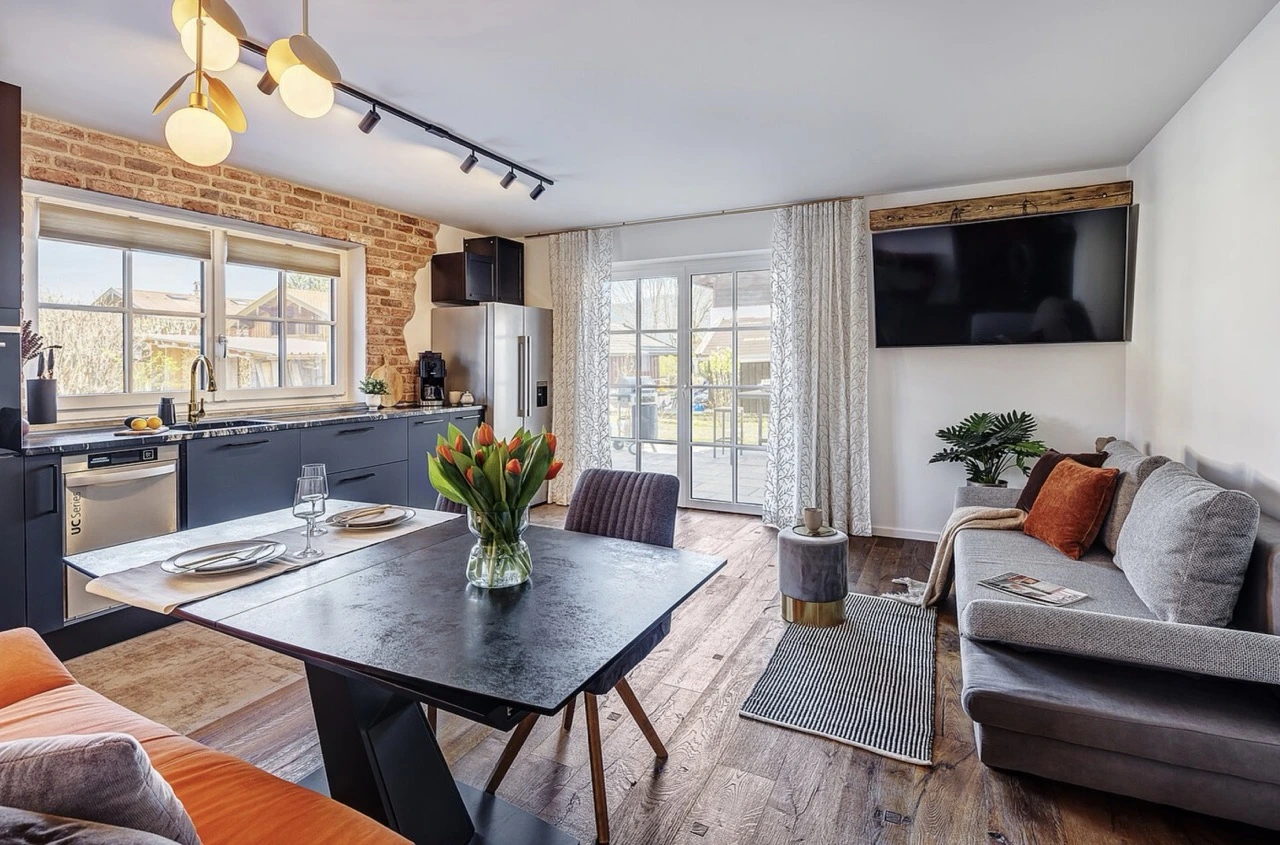 Modernes Wohnzimmer mit offener Küche, Esstisch mit Tulpenstrauß und Sofa-Ecke vor heller Fensterfront