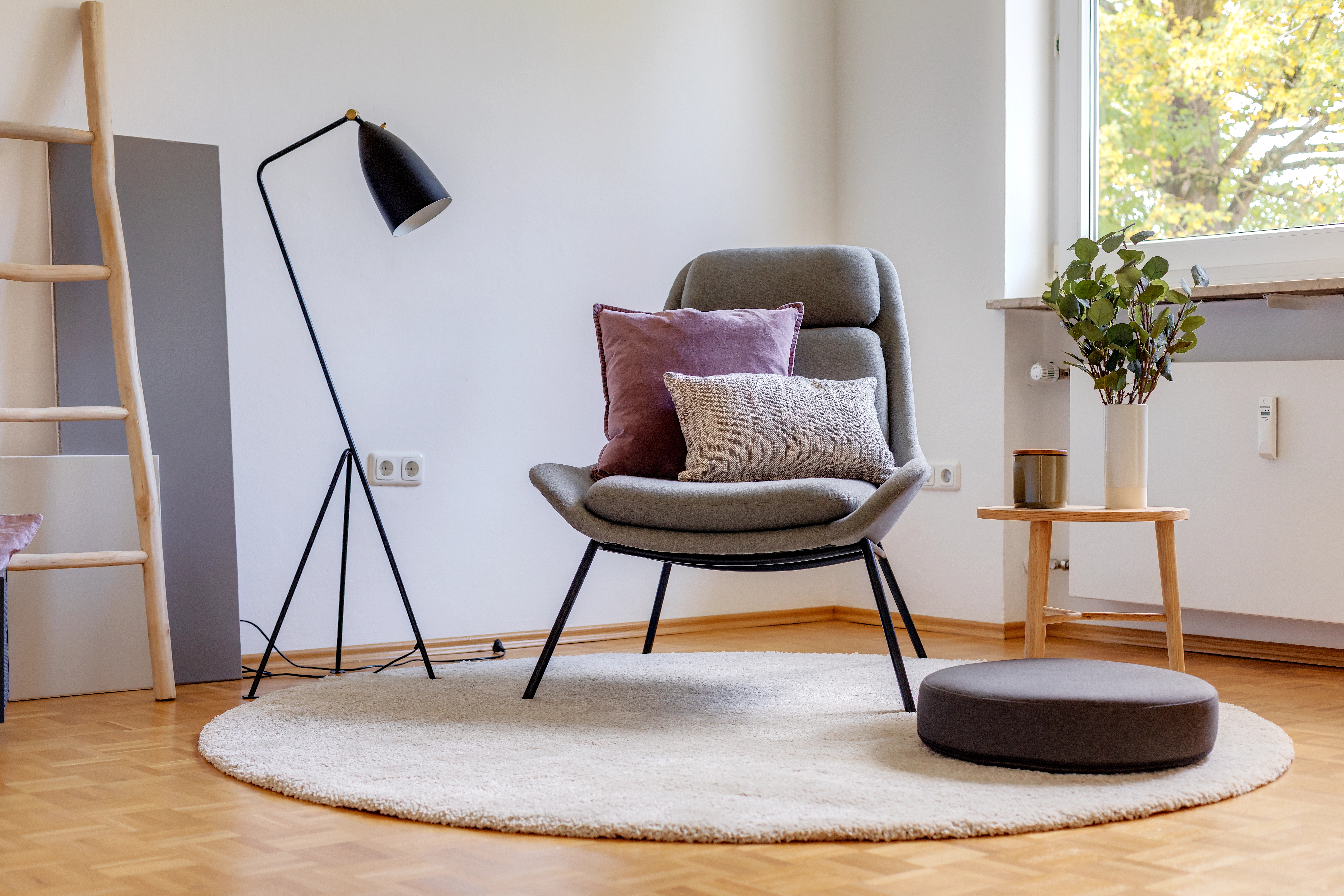 Hochwertig eingerichtetes Wohnzimmer mit grauem Designsessel, Stehlampe, kleinem Beistelltisch und rundem Teppich vor heller Wand