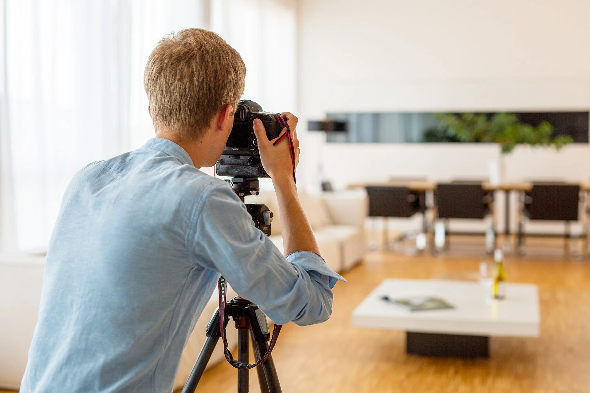 Fotograf mit Kamera auf Stativ fotografiert ein hell eingerichtetes Wohnzimmer mit Sofa und Couchtisch